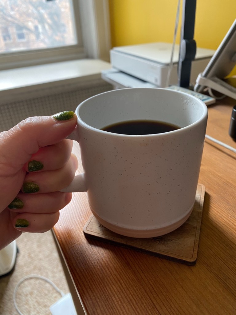 A hand with sparkly green nail polish holds a small white ceramic coffee mug by its handle. The body of the mug sits on a coaster on a brown desk in front of a yellow painted wall and a window with daylight coming in.